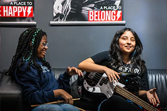 Two Performance Program Students Sitting Near One Another Holding Drumsticks and a Guitar Before Band Rehearsal