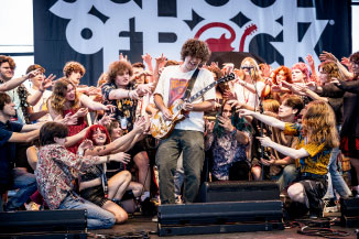 A student guitarist performing on stage, surrounded by peers who cheer him on.