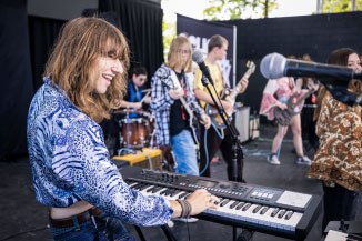 A student plays the keyboard, grinning on stage alongside peers while overlooking the audience.