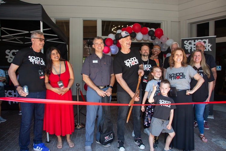 A group of people gathered at the entrance to the School of Rock in Pleasanton.  One is holding a large pair of ceremonial scissors and preparing the cut the grand opening ribbon.