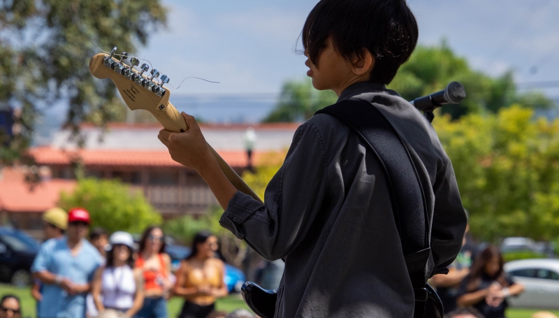 A student plays guitar at an outdoor concert, view from behind the stage.