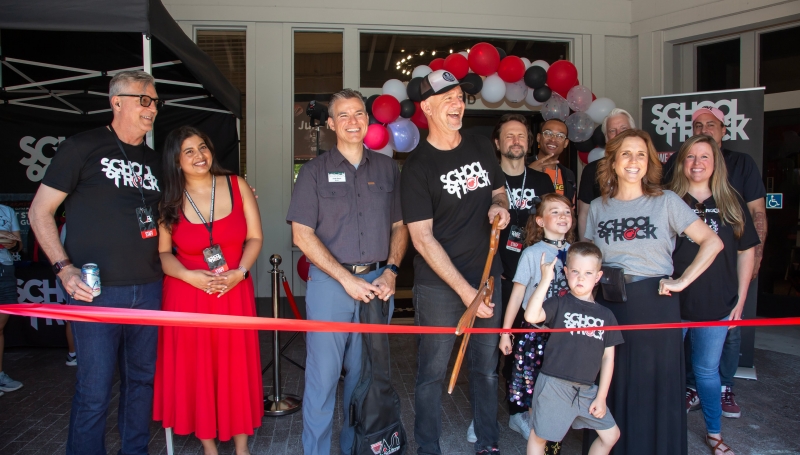 A group of people gathered at the entrance to the School of Rock in Pleasanton.  One is holding a large pair of ceremonial scissors and preparing the cut the grand opening ribbon.